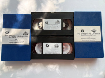 Two vintage VHS tapes with blue jewel cases are stacked vertically on a white surface with soft leaf shadows. The center tape is open, showing the black plastic tape reels inside, and both tapes feature white labels with text and a BMW logo.