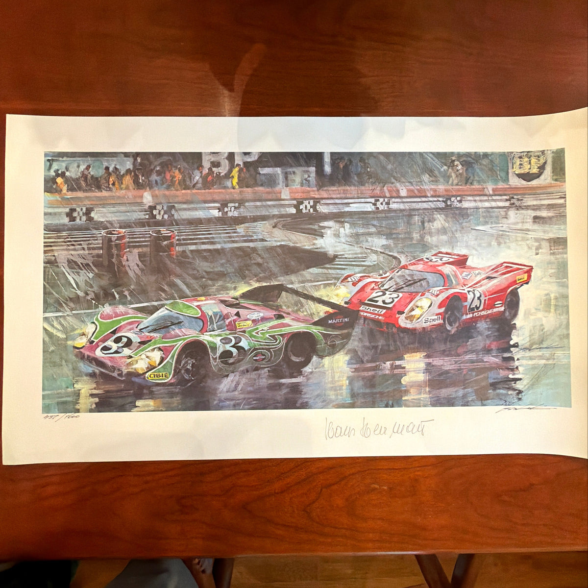 A vertical art print featuring two race cars on a racetrack, displayed on a wooden table. The top car is green and pink with the number 3, and the bottom car is red with the number 55.