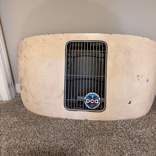 A vintage beige car grille with a black mesh center section and a circular blue, red, and white PCA emblem, resting on a carpeted floor against a plain wall.