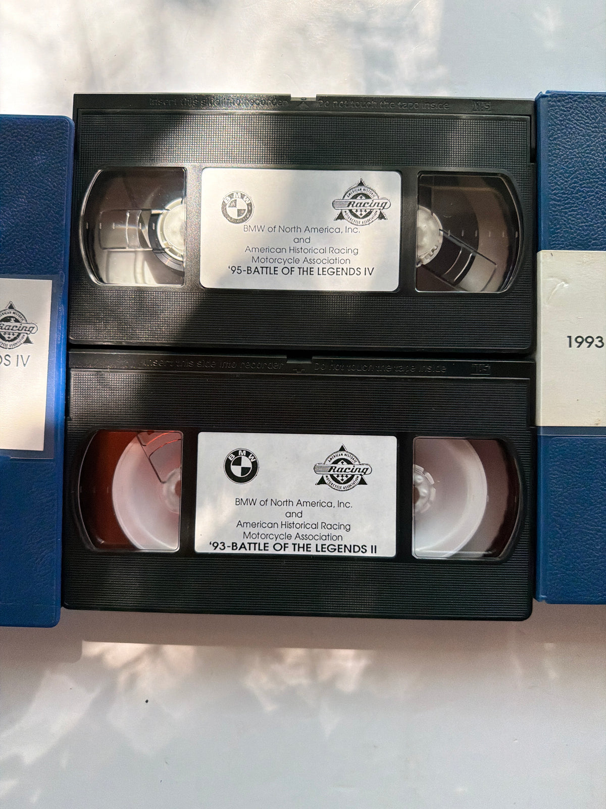 Two black VHS tapes from 1993 for BMW Historical Racing stacked vertically, with blue protective cases partially visible on both sides on a light surface.