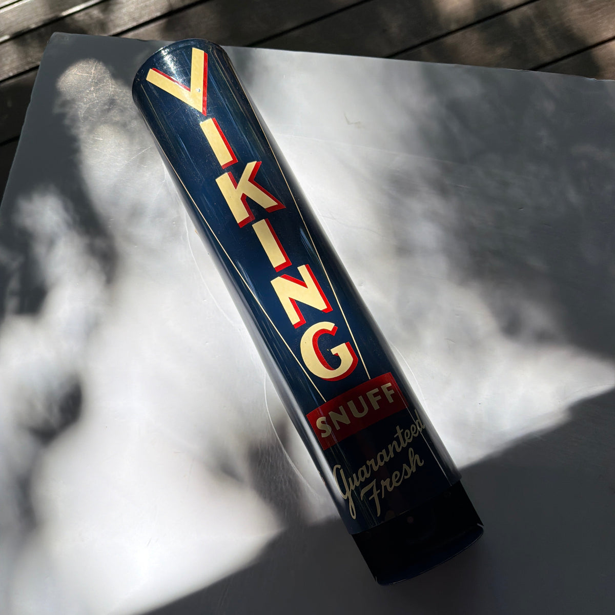 A vintage cylindrical metal sign for 'Viking Snuff' with cream lettering outlined in red on a dark blue background, photographed outdoors on a white surface with dappled sunlight and shadows.